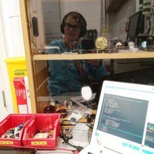 A photo of a laptop with computer code on it in front of a cupboard full of optical components