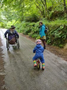 3 people walking in the woods. One is in a wheelchair, another is a toddler on a small bike, the third on foot.