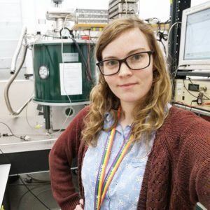 A selfie of a white woman with brown hair wearing glasses. Behind her is a superconducting magnet on an optical table