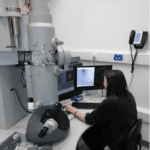 Caroline sitting with her hands on the controls of an a tall gray cylindrical machine which is an electron microscope