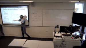 A picture of Gwen, a blonde white fairly tall woman, in the front of a classroom, pointing at a projector screen to explain statistics. 